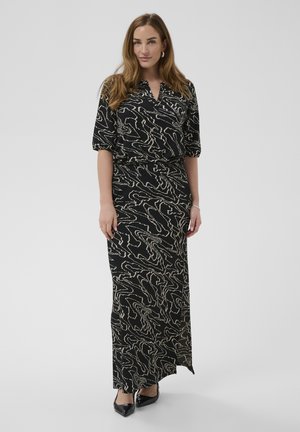 Žena s dlhými vlasmi, oblečená v čiernych a béžových šatách s abstraktným vzorom, s rukávmi po lakte a čiernymi špicatými topánkami, stojaca na jednoduchom pozadí.