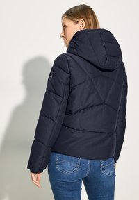 Veste rembourrée navy avec capuche, présentant des panneaux diagonaux matelassés et une finition texturée. Détail de badge attaché sur la manche gauche.