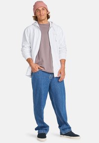 Sudadera gris claro con cremallera sobre una camiseta topo, combinada con vaqueros azules holgados. Un gorro de punto rosa y zapatillas oscuras completan el look.
