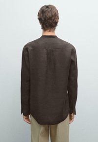 Brown long-sleeve shirt with a straight cut, back pleat detail, soft texture, and buttoned cuffs, worn over light-colored trousers.