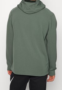 Personne portant un sweat à capuche côtelé vert olive et un pantalon noir, debout de dos, devant un fond uni.