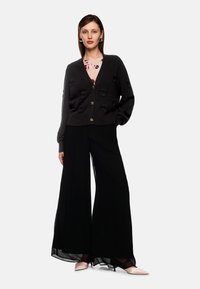 Schwarze Strickjacke mit floralen Details und goldenen Knöpfen, die über einem rosa Blumenshirt getragen wird. Kombiniert mit weiten schwarzen Hosen und blassrosa Schuhen.