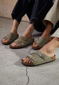 Žalios suede šlepetės su dviem reguliuojamais sagtimis, kamštinė padas ir tekstūruota guma, pateikiamos ant kojų prieš betoninę paviršių.