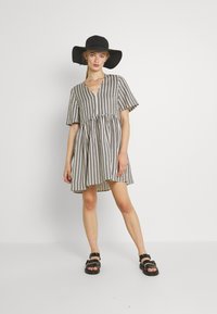 Gestreiftes kurzes Kleid in Beige und Schwarz, mit V-Ausschnitt, kurzen Ärmeln, schwingendem Design und gerafftem Taillenbereich; kombiniert mit schwarzen Sandalen und einem Hut.