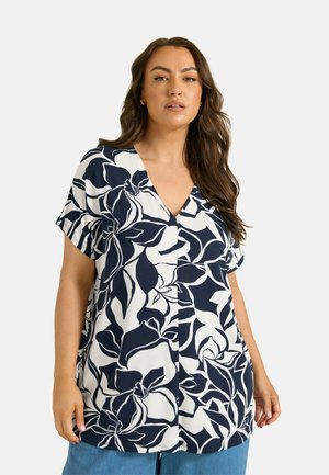 Kvinde med langt brunt hår iført en hvid bluse med stort sort blomsterprint og blå bukser mod en ensfarvet baggrund.