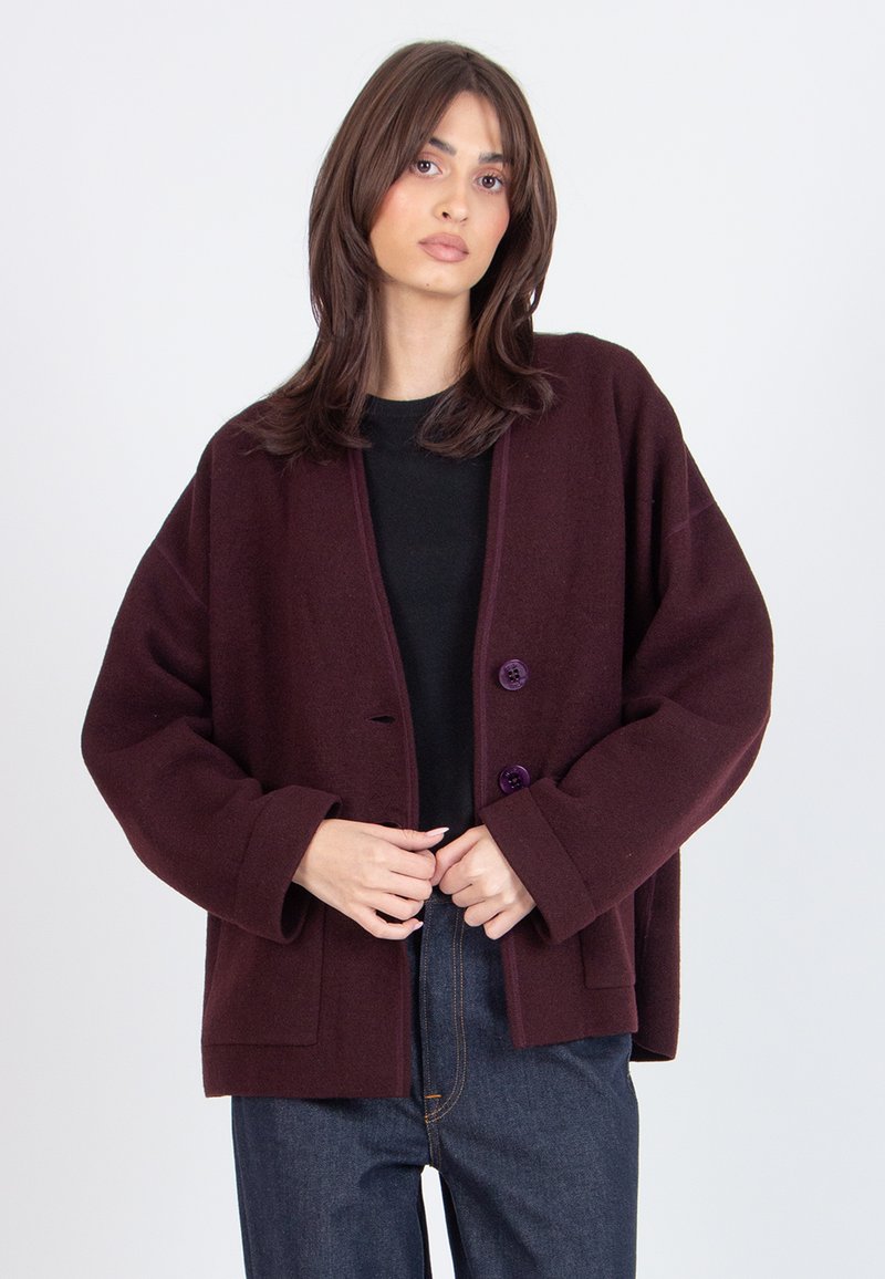 Woman with shoulder-length brown hair wearing a dark purple oversized buttoned jacket over a black top and blue jeans.