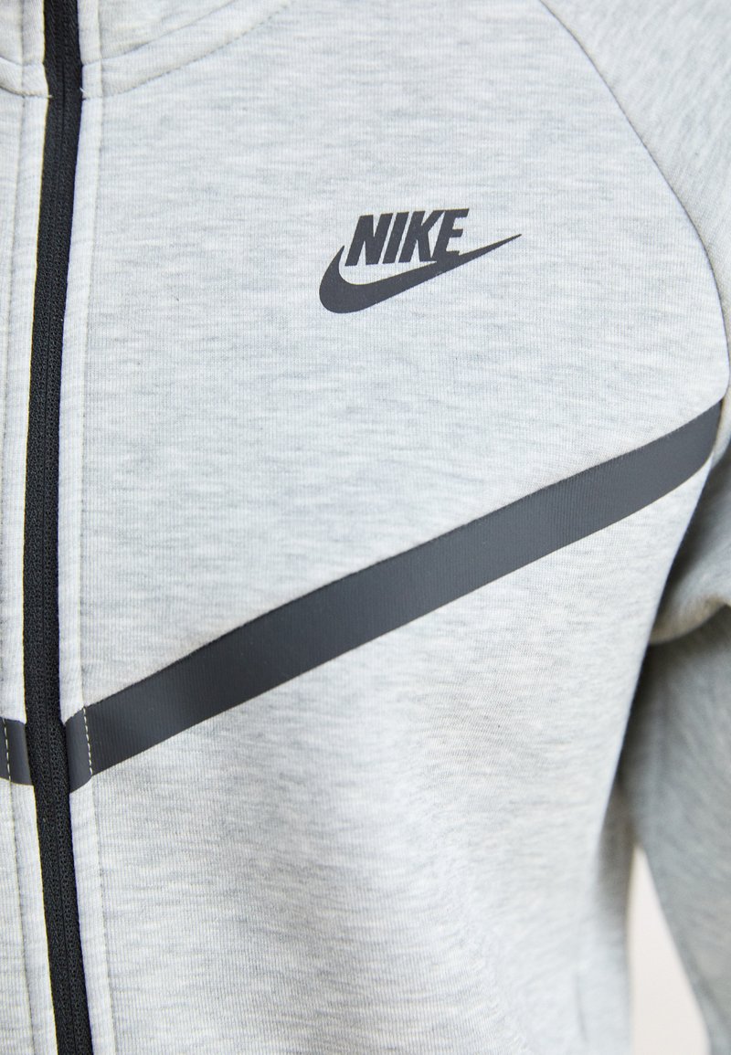 Lynlåsjakke i lysegrå sweatshirtstof med et sort Nike-logo og vandrette sorte accentstriber over brystet.