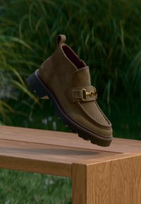 Mocassin en daim marron avec boucle dorée et semelle noire rainurée flottant au-dessus d'un banc en bois en plein air avec un arrière-plan de feuillage vert.