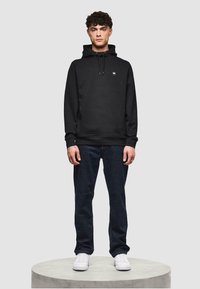 Schwarzer Hoodie mit Kordelzugkapuze, Fronttasche und kleinem Logodetail. Kombiniert mit dunkelblauen Jeans und weißen Sneakers.