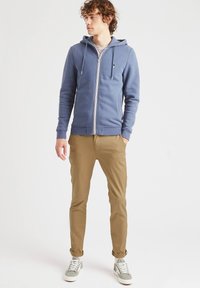 Felpa blu con zip, cappuccio regolabile, polsini a coste e tasche, abbinata a pantaloni slim-fit beige e sneakers grigie.