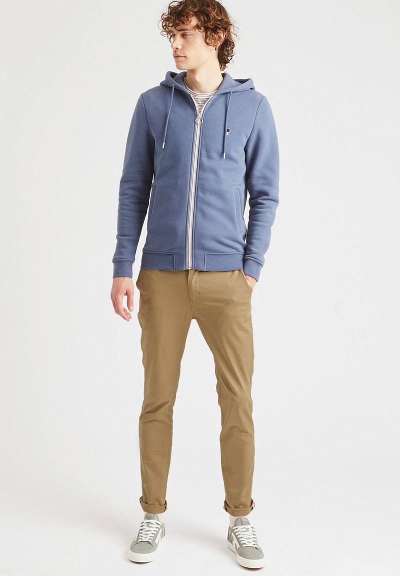 Felpa blu con zip, cappuccio regolabile, polsini a coste e tasche, abbinata a pantaloni slim-fit beige e sneakers grigie.