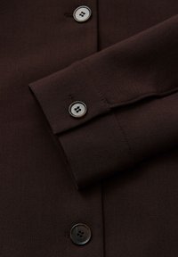 Bruine mouw van een shirt met een gladde stof, dubbele manchet met knopen en grijze knopen, met strakke stiksels en een gestructureerd ontwerp.