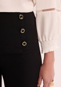 Czarne spodnie z trzema złotymi guzikami z boku, połączone z białą, miękką bluzką z detalem elastycznych mankietów.