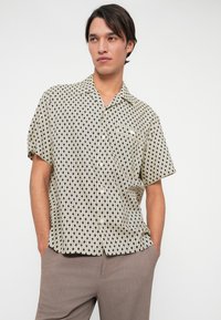 Korte mouwen overhemd in crème met een zwart geometrisch patroon. Voorzien van een puntige kraag en een enkele borstzak. Lichtgewicht stof.