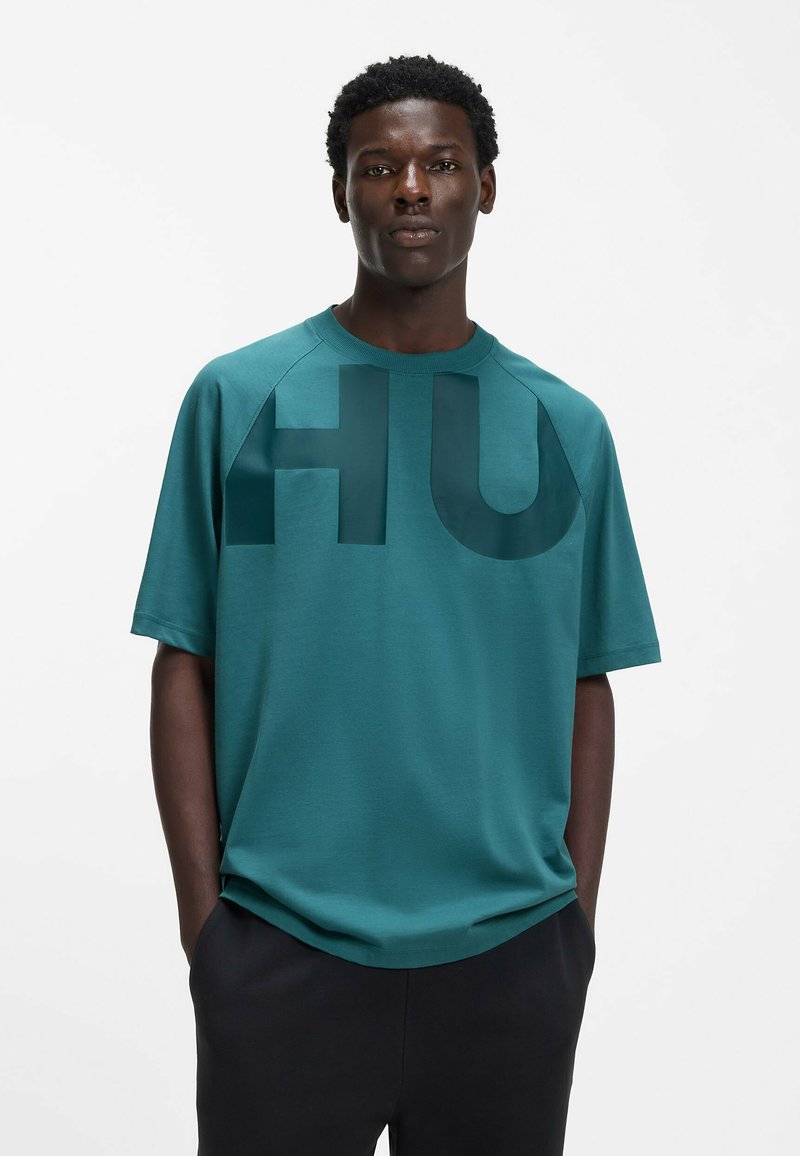 Krótki rękaw t-shirt w kolorze morskim, wykonany z gładkiej tkaniny. Na przodzie znajduje się duży, ciemny nadruk "HU". Klasyczny, okrągły dekolt.