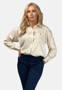 Kremowa satynowa bluzka z kieszenią na piersi, złotymi guzikami i luźnym krojem, zestawiona z granatowymi spodniami z bocznymi kieszeniami i złotymi akcentami.