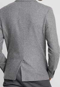 Man i en välsittande grå kavaj med mittbaksvep och matchande grå byxor, stående mot en helt vit bakgrund.