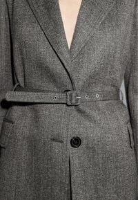 Grau strukturierter Blazer mit Reverskragen, einem einzelnen schwarzen Knopf und einem passenden Stoffgürtel, der in der Taille befestigt ist.