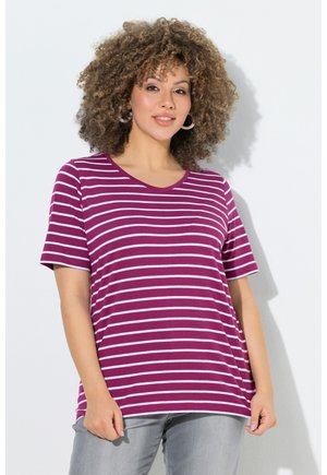 Woman with curly hair wearing hoop earrings, a purple and white striped short-sleeve shirt, and light gray pants, standing against a white wall.