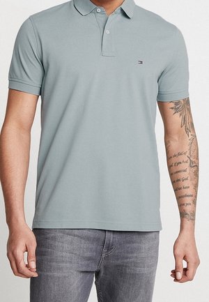 Mężczyzna ubrany w jasnoszarą koszulkę polo z krótkim rękawem i szare dżinsy, z widocznym tatuażem na prawym przedramieniu, przedstawiającym tekst i wzór kwiatowy.