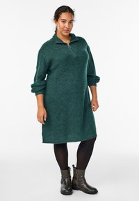 Žalias megztas suknelė su pusiau užtraukiamu kaklu ir sulankstytais rankogaliais, derinama su juodomis pėdkelnėmis ir kulkšnies aulinukais. Tekstūruoto, laisvo silueto dizainas.