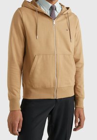 Bruine rits hoodie gemaakt van een zachte stof, voorzien van een capuchon, kangoeroe zakken, geribbelde manchetten en een klein logo op de borst.