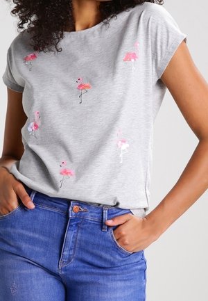 Mujer con una camiseta gris claro con pequeños estampados de flamencos rosas, combinada con vaqueros azules de mezclilla, manos en los bolsillos.