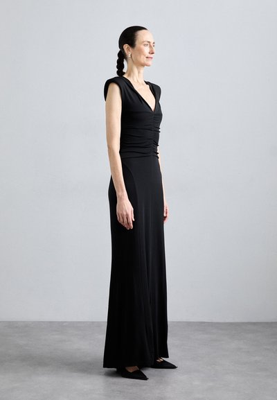 Sort kjole med tætsiddende overdel, v-hals og korte ærmer. Stoffet er glat og falder elegant til gulvet. Båret med spidse flade sko.