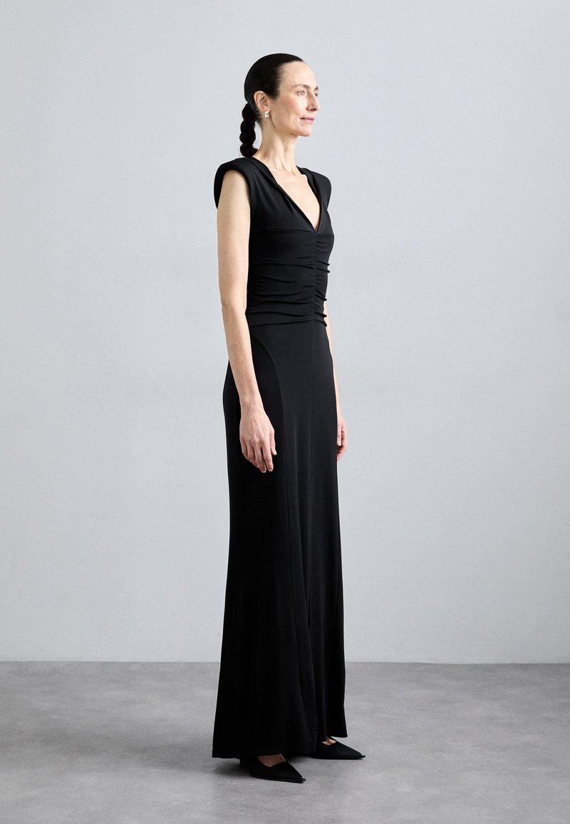 Vestido negro con un corsé ajustado, escote en forma de V y mangas cortas. La tela es suave y cae elegantemente hasta el suelo. Combinado con zapatos planos de punta.