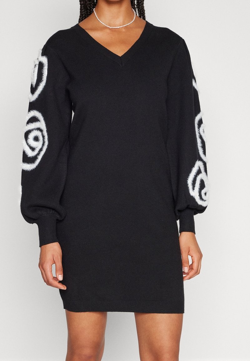 Robe-pull noire à col en V avec de longues manches bouffantes, présentant des motifs spirales blancs et duveteux sur les manches et un ourlet court.