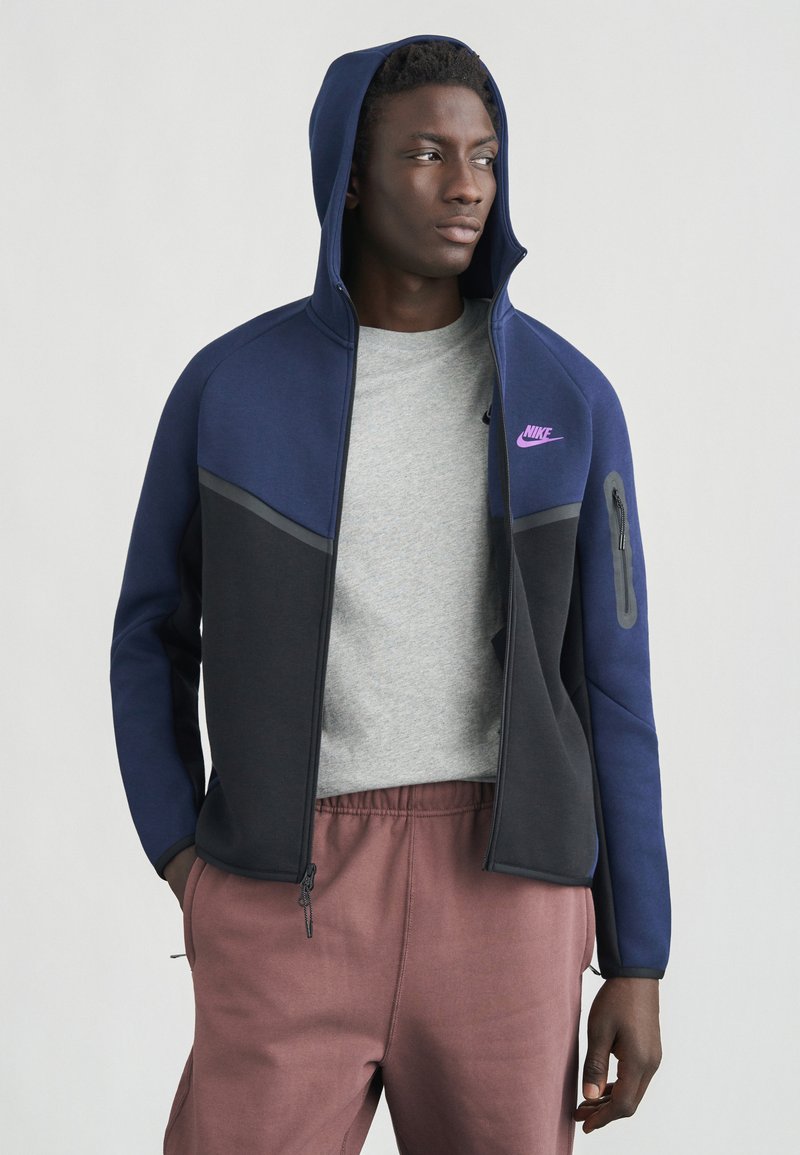 Tumši zils un pelēks džemperis ar rāvējslēdzēju priekšpusē, divtoņu dizains, violets logotips krūtīs un sānu rāvējslēdzēja kabata. Valkāts virs pelēkas krekla.