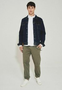Jeansowa kurtka w ciemnoniebieskim kolorze, biała koszula zapinana na guziki, oliwkowe spodnie i białe sneakersy. Prosty, czysty design z funkcjonalnymi detalami.
