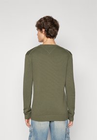 Pull en tricot côtelé vert olive à manches longues et col rond. Il présente une finition texturée et des poignets ajustés. Porté avec un jean bleu clair.