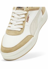 Gros plan sur une basket Puma beige et blanche avec des lacets blancs et un logo sur le côté et la languette.