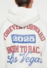Sweatshirt branco com uma grande impressão gráfica nas costas. Texto em vermelho e azul que diz "PARA SEMPRE AVANTE 2025 NASCIDO PARA CORRER Las Vegas." Material suave.