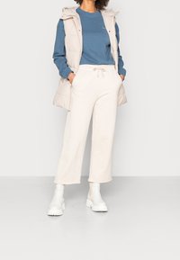 Chaleco acolchado beige, camisa de manga larga azul y pantalones deportivos anchos color crema, combinados con botas blancas hasta el tobillo, sobre un fondo claro.