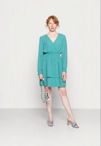Vestido envolvente azul-petróleo com mangas longas e cintura elástica, com barra em camadas, combinado com uma mala prateada e sandálias de salto xadrez.