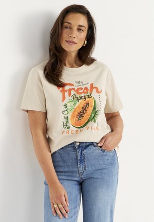 Kvinde iført beige t-shirt med papaya-grafik og teksten "Fresh Papayas Fruit Club Fresh Vibes", kombineret med blå jeans, stående mod en hvid væg.