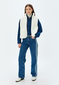 Weißer Fleece-Weste über einer dunkelblauen Jeansjacke und hellblauen Jeans mit seitlichem Streifen. Weiße Stiefeletten runden den Look ab. Saubere, schlichte Linien.