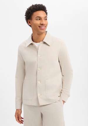 Jeune homme souriant, portant une veste beige boutonnée avec col et un pantalon assorti, debout avec une main dans la poche.