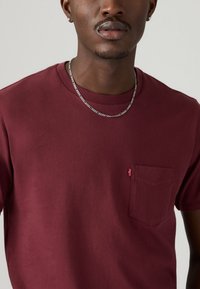 Bordeaux Baumwoll-T-Shirt mit Rundhalsausschnitt, kurzen Ärmeln und einer Brusttasche, die mit einem kleinen roten Markenetikett versehen ist. Glatte Textur.