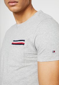 Graues Baumwoll-T-Shirt mit einer Brusttasche, die marineblaue, rote und weiße Streifen aufweist. Kurze Ärmel mit einem kleinen Markenlogo am Bund.