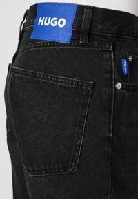 Acercamiento a un bolsillo trasero de unos vaqueros de mezclilla negros con etiqueta y etiqueta azul que presenta la marca blanca "HUGO".