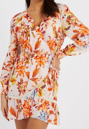 Kobieta ubrana w długą sukienkę kopertową z długim rękawem, ozdobioną pomarańczowo-czerwono-żółtymi kwiatowymi wzorami i koronkowym wykończeniem, z ręką opartą na biodrze.