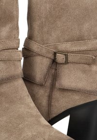 Botas de tobillo de ante color marrón claro con una correa decorativa y una hebilla metálica rectangular, además de un tacón de aguja. Textura suave y diseño minimalista.