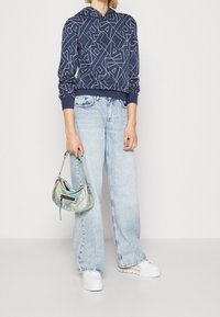 Felpa navy con motivo geometrico bianco, abbinata a jeans a gamba larga blu chiaro e una borsa iridescente, che mette in mostra texture lucide.