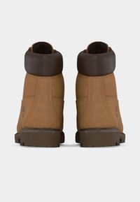 Ζευγάρι καφέ δερμάτινων μποτάκια αστραγάλου με σκούρα επενδεδυμένα περιλαίμια και υφασμένα πέλματα, εμφανιζόμενα από πίσω σε λευκό φόντο.