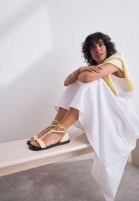 Kollased rihmikud sandaalid minimalistliku disainiga ja musta tasapinnalise tallaga. Rihmad on varustatud pahkluu pandlaga ning neil on sile ja matt tekstuur.