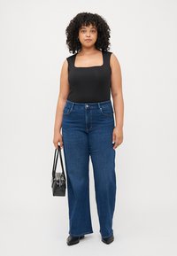 Black square-neck bodysuit, blue wide-leg jeans, and a small black handbag. Curly hair and neutral background. Smooth fabric and classic cut.