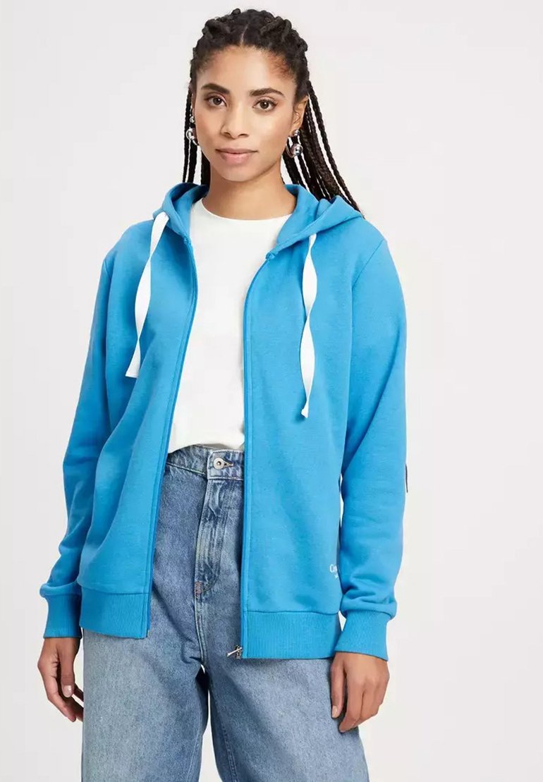 Niebieska bluza z kapturem na zamek, wykonana z miękkiego materiału, z przednim zamkiem, białymi sznurkami i ściągaczami na rękawach. W połączeniu z białą koszulką i dżinsami.
